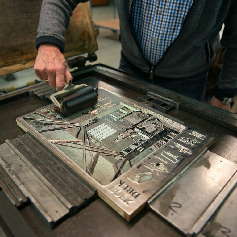 Person rollt Druckfarbe über eine Druckplatte mit verschiedenen Buchstaben und Formen.